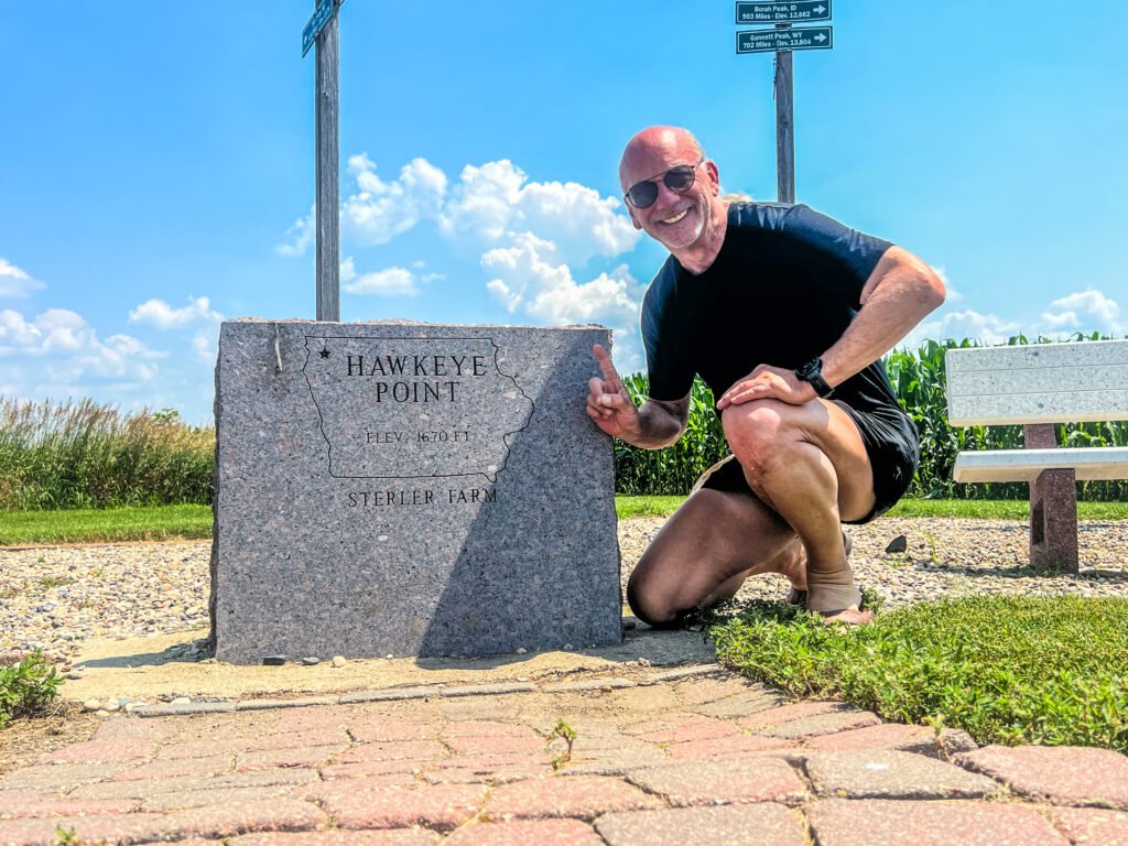 Stephen kneeling beside the Hawkeye Point granite marker reading 1,670 feet elevation on Sterler Farm in Iowa.