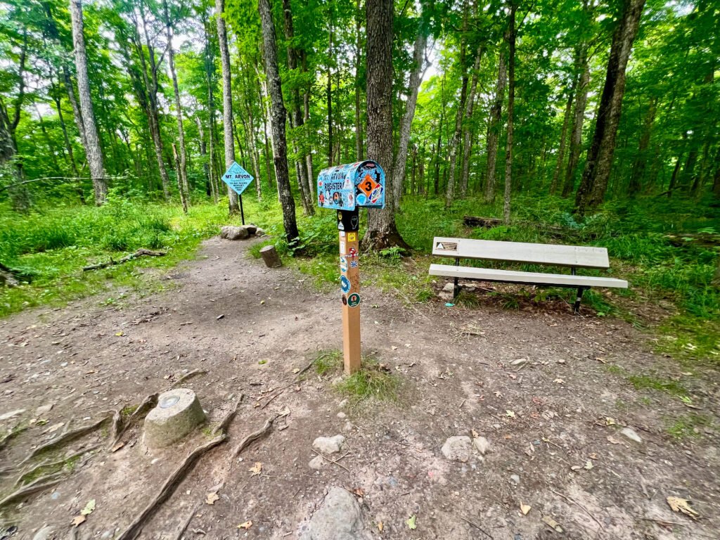 Mount Arvon summit area featuring the Highpointers mailbox register, benchmark marker, and memorial bench in Michigan’s Upper Peninsula forest.