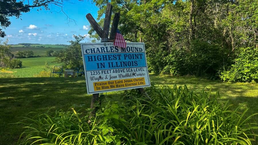 Welcome sign at Charles Mound, highest point in Illinois at 1,235 feet, with American flag and farmland views in the background.