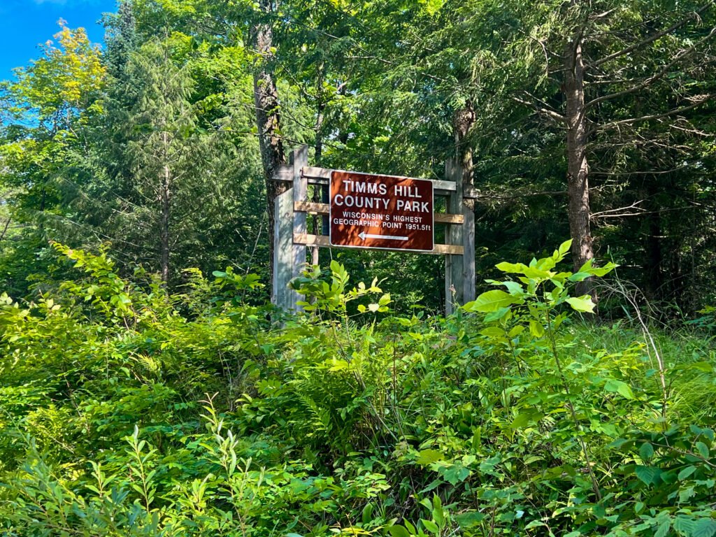 Timms Hill County Park entrance sign along County Road RR in Wisconsin surrounded by forest.