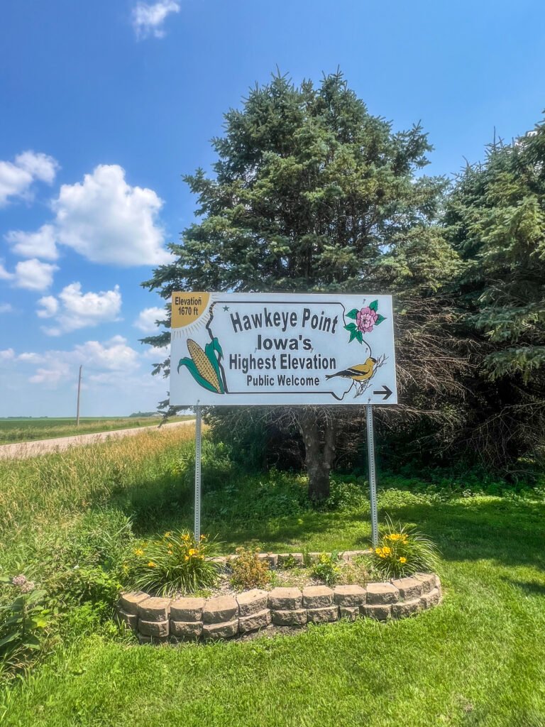 Hawkeye Point roadside sign on Iowa Highway 60 marking Iowa’s highest elevation at 1,670 feet with cornfields and trees behind it.