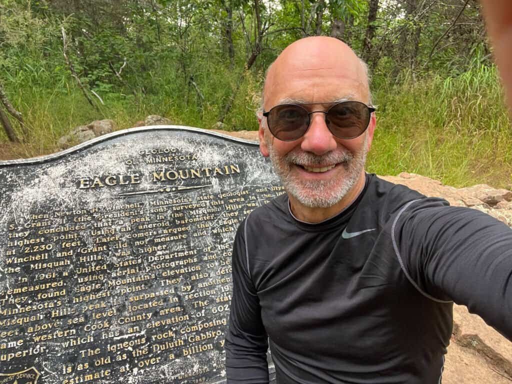 Smiling hiker taking a selfie at the Eagle Mountain summit plaque in Minnesota