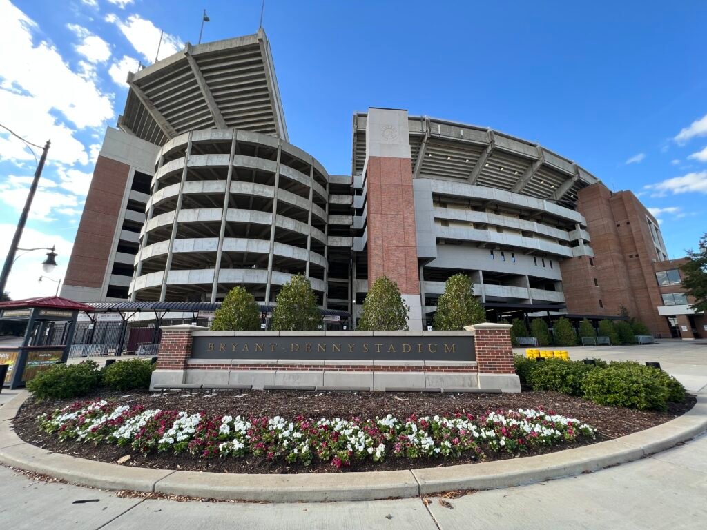 Bryant-Denny Stadium exterior at the University of Alabama in Tuscaloosa with landscaped entrance and blue sky overhead.