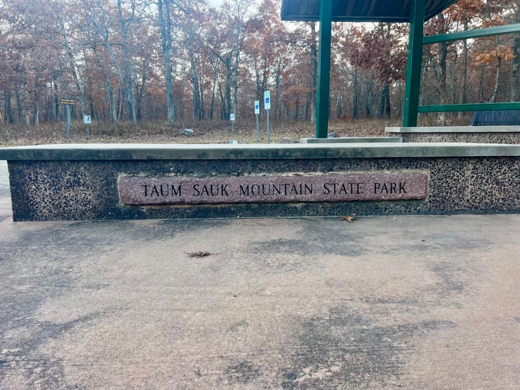 Stone sign for Taum Sauk Mountain State Park near paved trailhead and accessible parking area in fall.