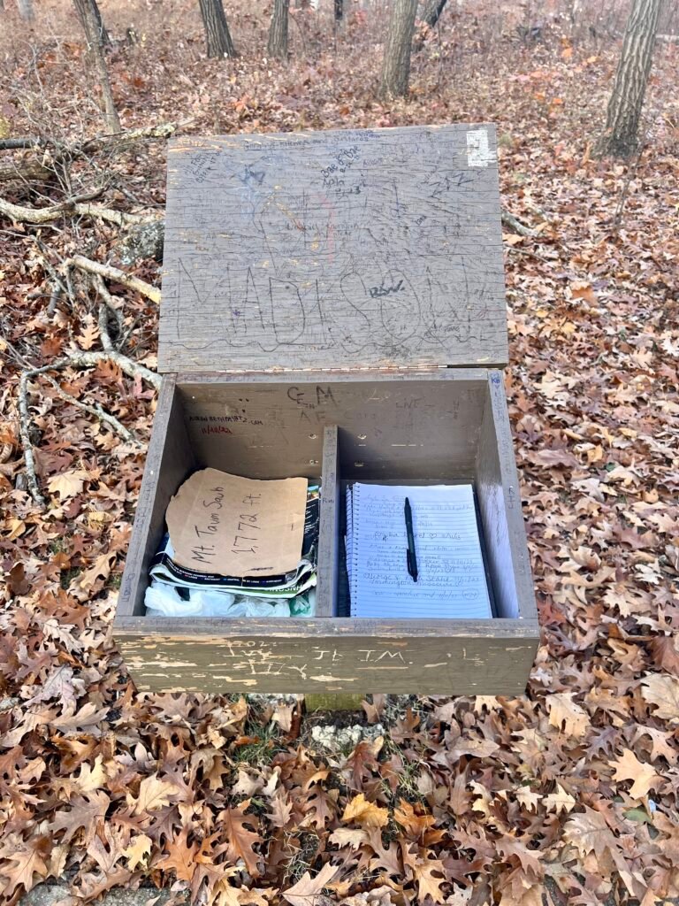 Wooden summit register box at Taum Sauk Mountain containing trail log book and pen among fallen leaves.