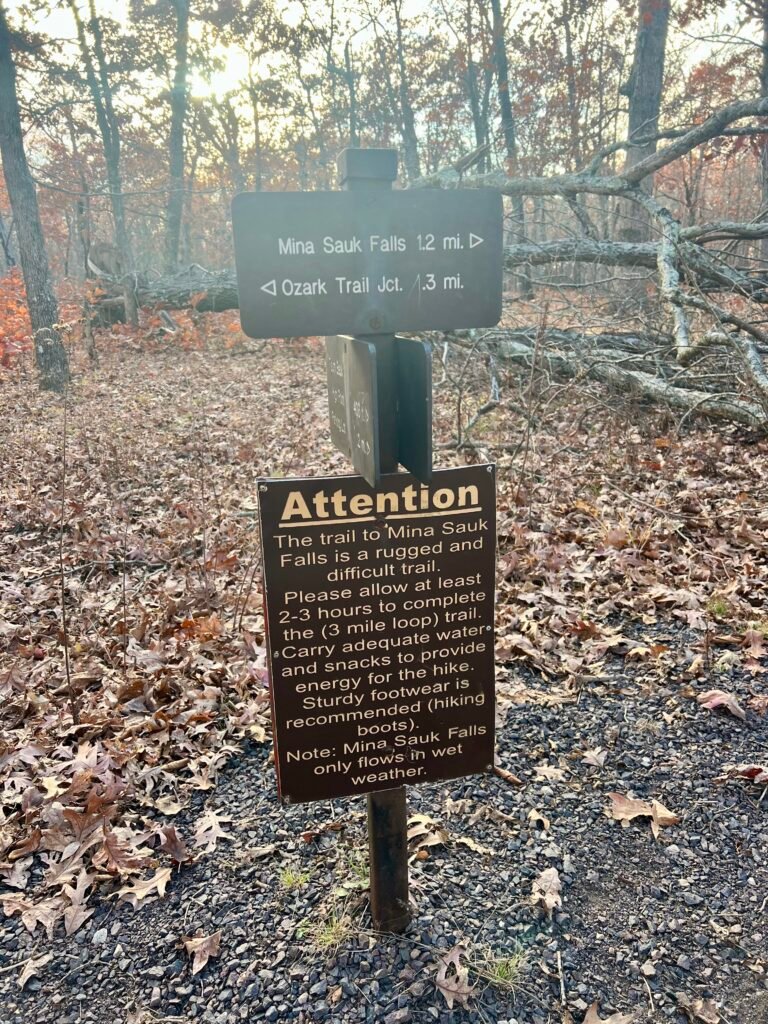 Trail sign for Mina Sauk Falls showing distances, difficulty warnings, and hiking recommendations at Taum Sauk Mountain State Park.
