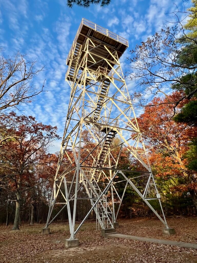 Steel lookout tower rising above fall foliage at Taum Sauk Mountain State Park under a bright blue sky.