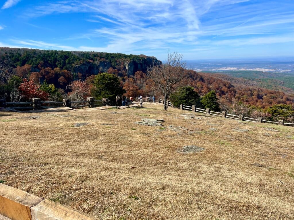 Wide view from Cameron Bluff Overlook’s first parking area, with stone fence, grassy lawn, and fall foliage across Mount Magazine State Park.