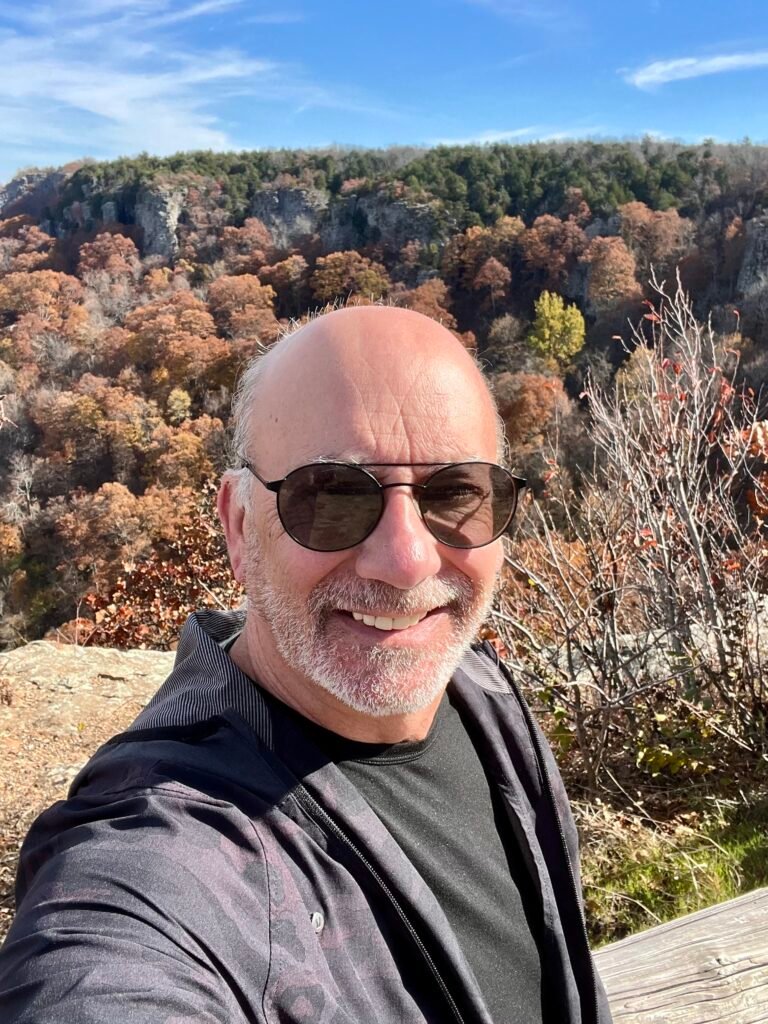 Smiling hiker at Mount Magazine State Park with fall foliage and rocky cliffs in the background.