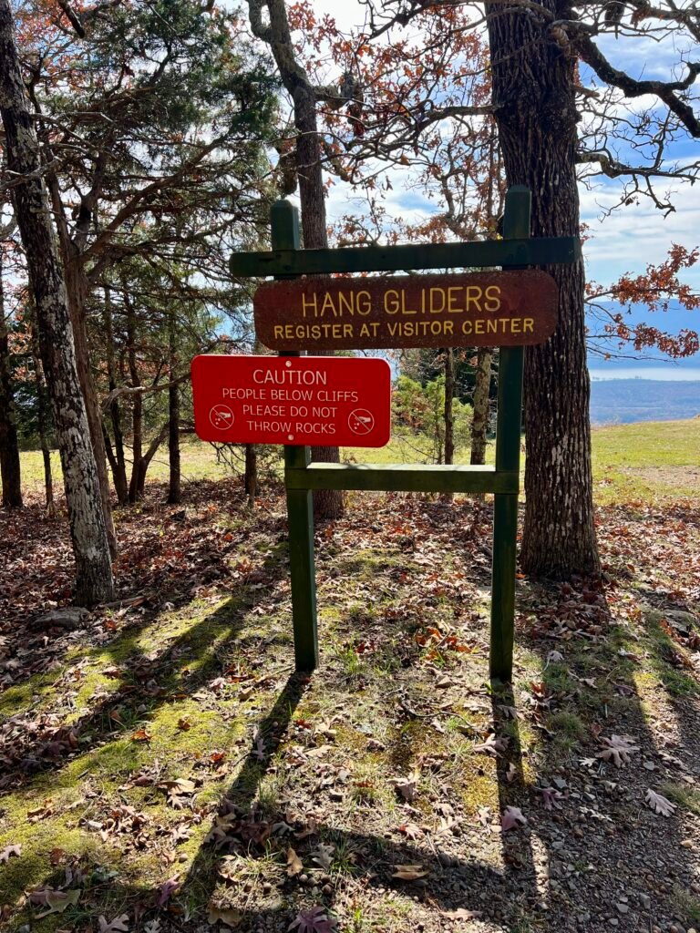 Wooden sign at Mount Magazine State Park marking the Hang Glider Launch Area, with a red caution sign warning not to throw rocks from the cliffs.