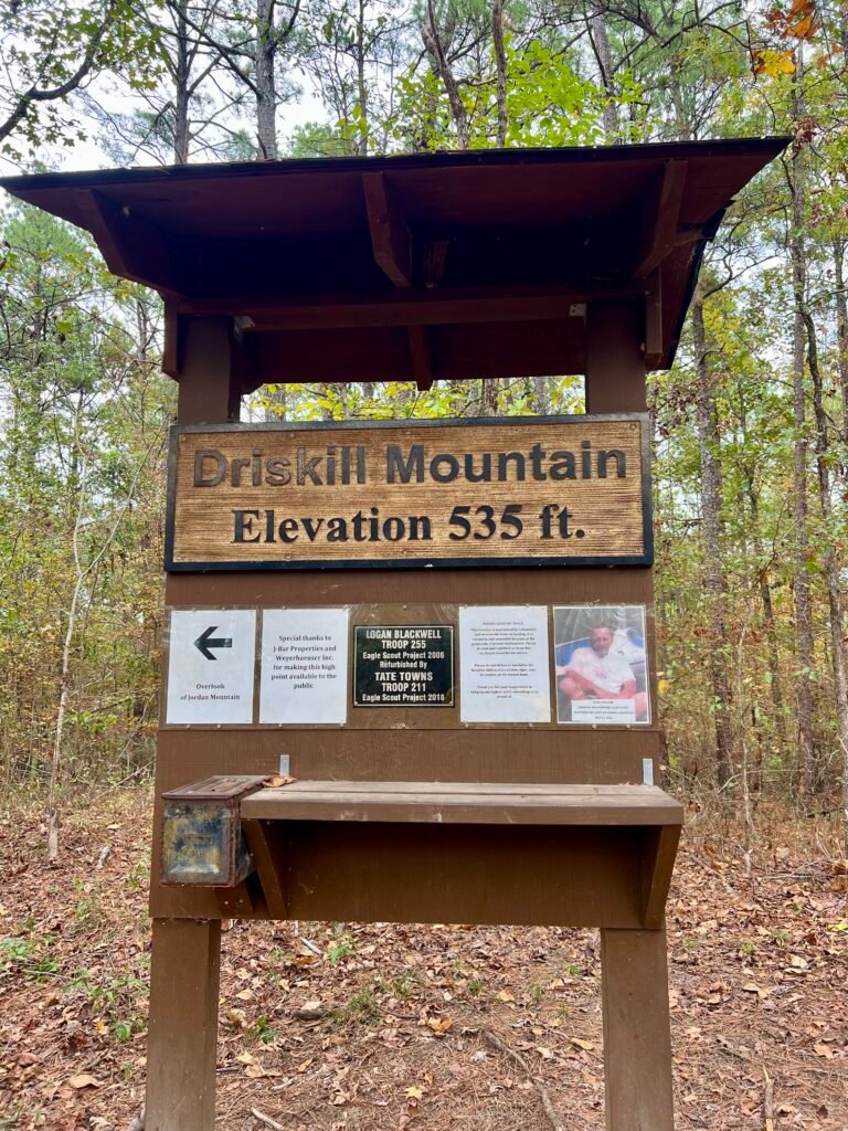 Wooden summit sign at Driskill Mountain showing elevation 535 feet, with a small ammo box on the left containing the highpoint registry.