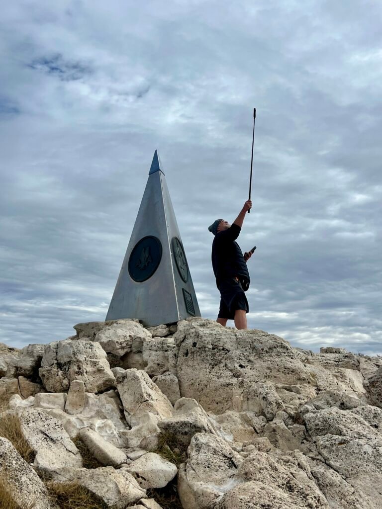Hiker standing beside the Guadalupe Peak monument using a selfie stick for a 360-degree summit photo