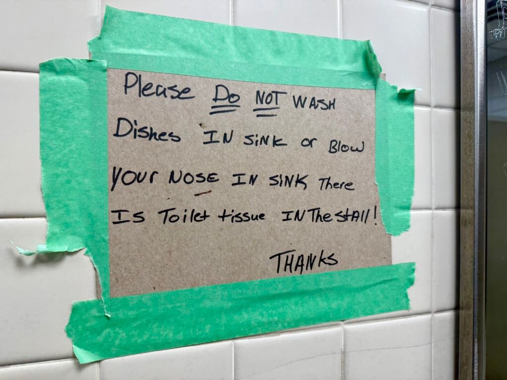 Handwritten sign taped to a tiled wall asking visitors not to wash dishes or blow their nose in the sink.