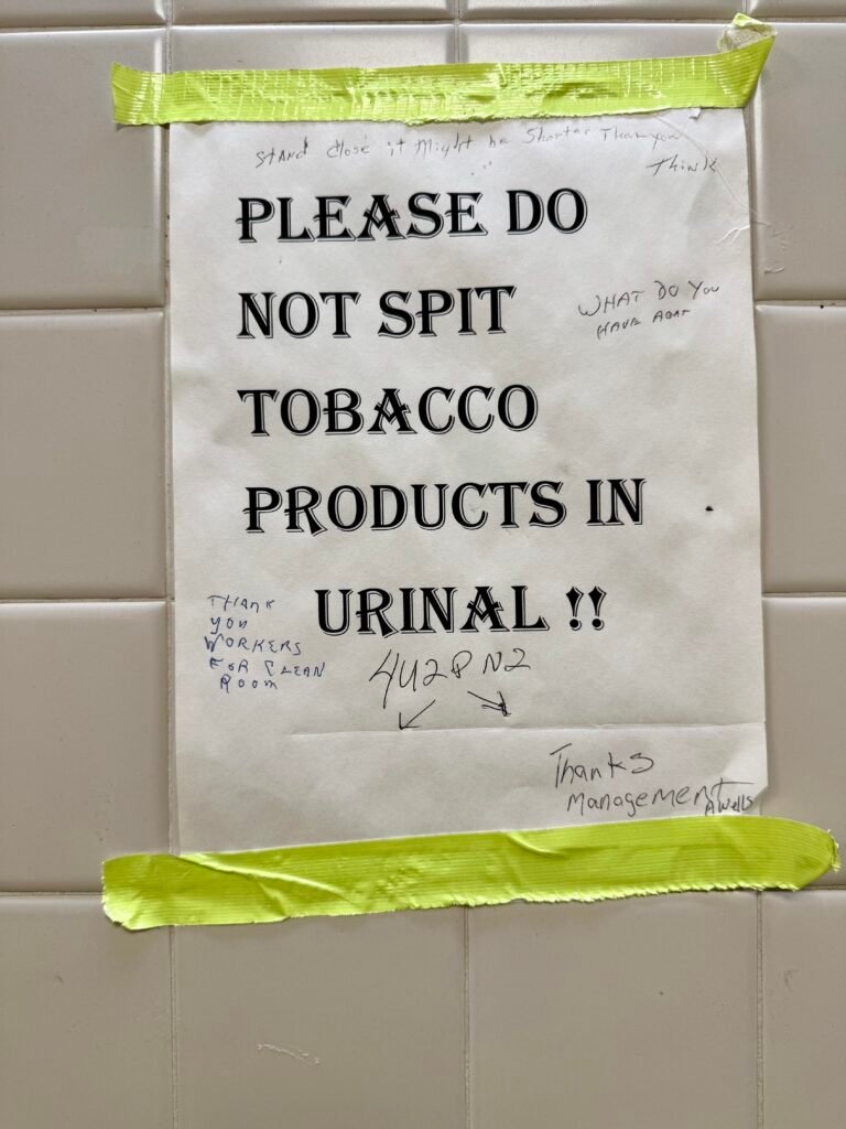 Printed restroom sign taped to a tiled wall requesting that tobacco products not be spit in the urinal, with handwritten notes added.