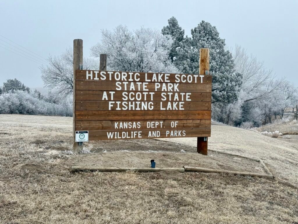 Entrance sign for Historic Lake Scott State Park in Kansas with frost-covered trees in winter.