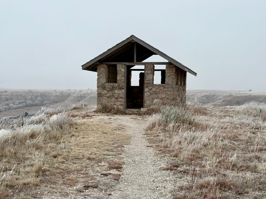 Small stone shelter building on the bluff at Historic Lake Scott State Park, surrounded by frost-covered prairie grass and overlooking the valley below.