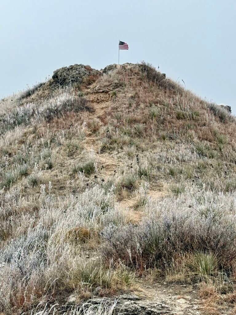 Steep dirt trail descending from the bluff top at Buzzard’s Point with frost-covered prairie grasses and an American flag visible above.