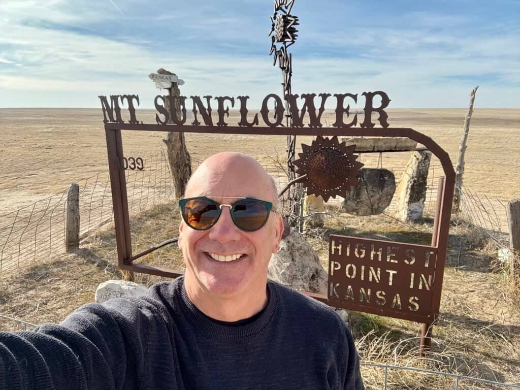 Smiling hiker taking a selfie at the Mount Sunflower Kansas high point with the summit sign and open plains behind.