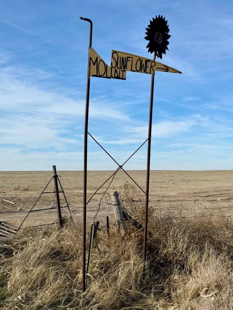 Rustic metal sign pointing toward Mount Sunflower with open prairie and wind turbines on the Kansas High Plains.