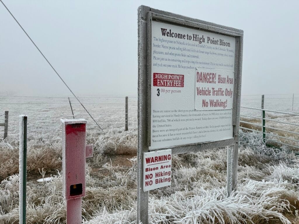 Red donation box and welcome sign at the entrance to High Point Bison Ranch near Panorama Point, Nebraska, coated in frost and surrounded by winter grass.