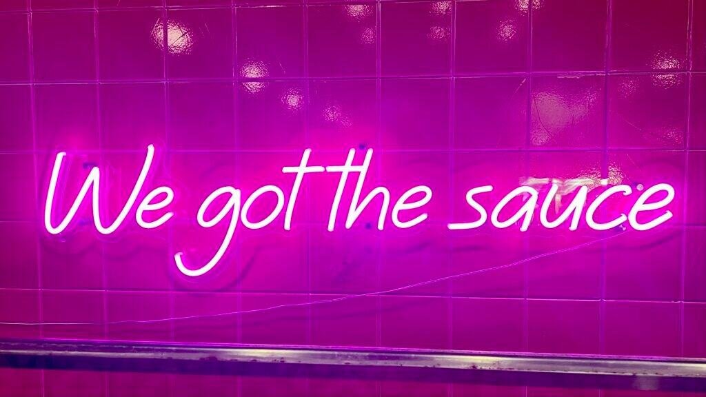 Neon sign reading “We got the sauce” glowing against a pink tiled wall