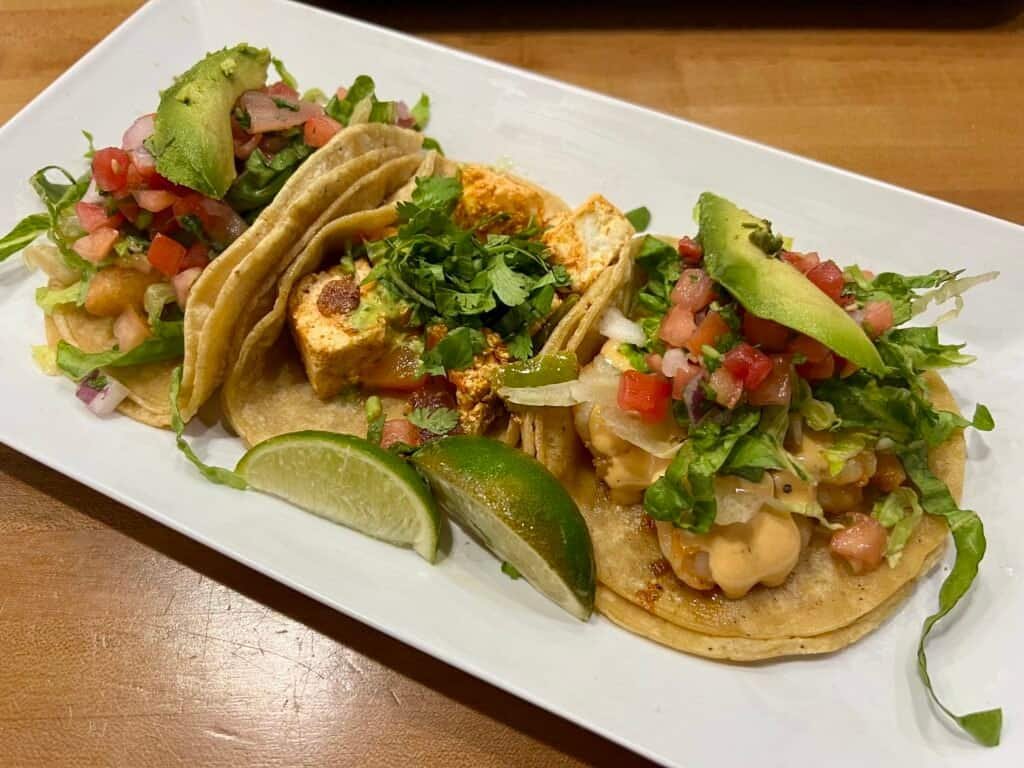 Trio of tacos topped with avocado, pico de gallo, and cilantro served with lime wedges on a white plate