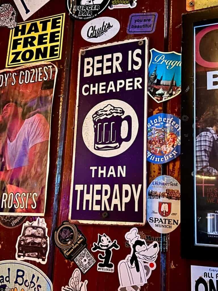 Sticker-covered wall at Rossi’s with a “Beer Is Cheaper Than Therapy” sign