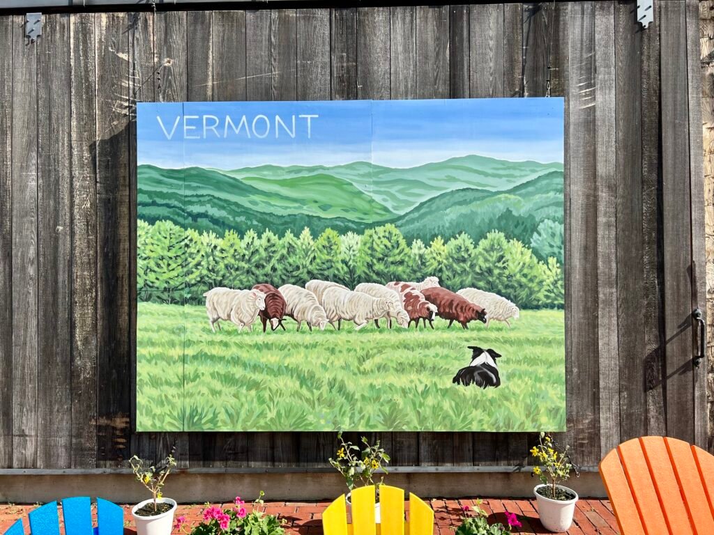 Painted barn mural of sheep grazing in a green Vermont field with mountains in the background.