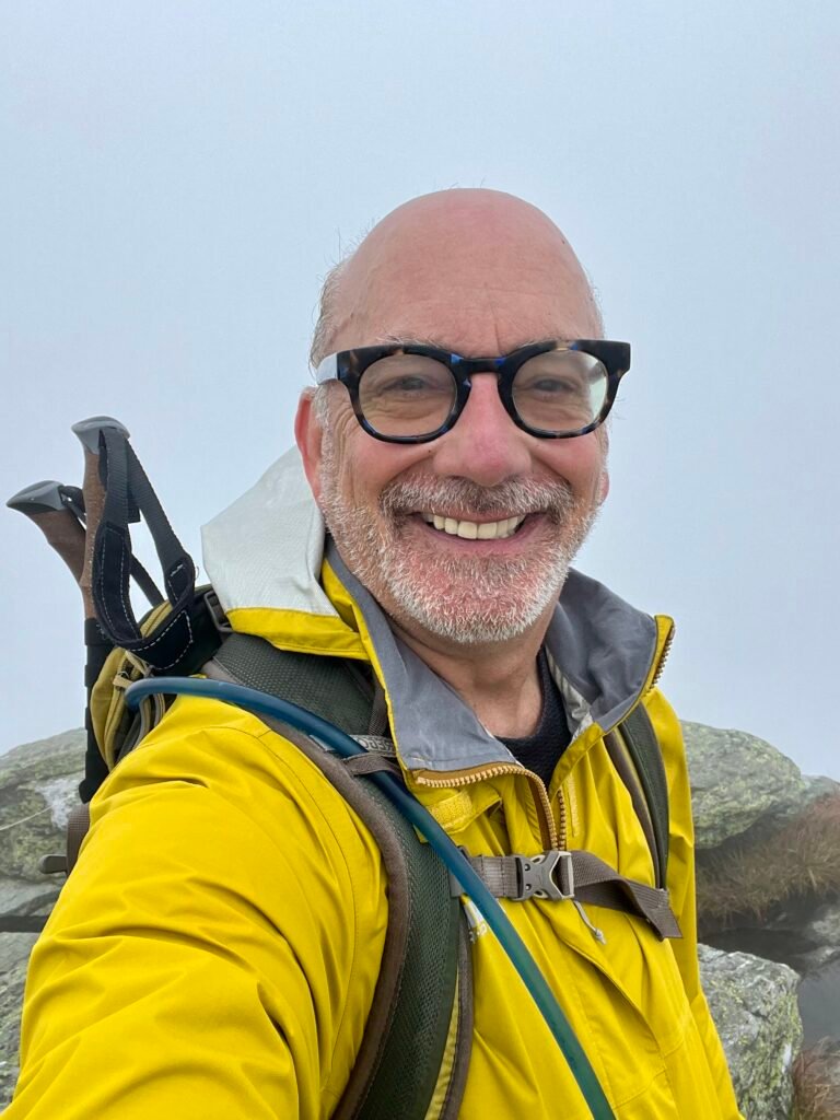 Hiker in yellow jacket smiling at the foggy Mount Mansfield summit.