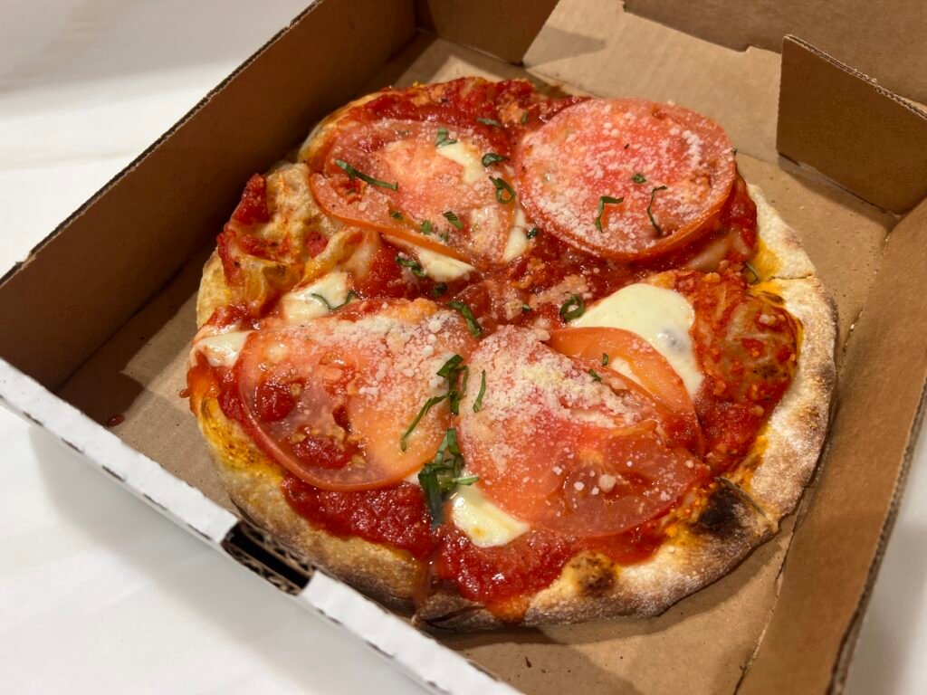 Personal-sized tomato and mozzarella pizza in a takeout box with fresh tomato slices on top.